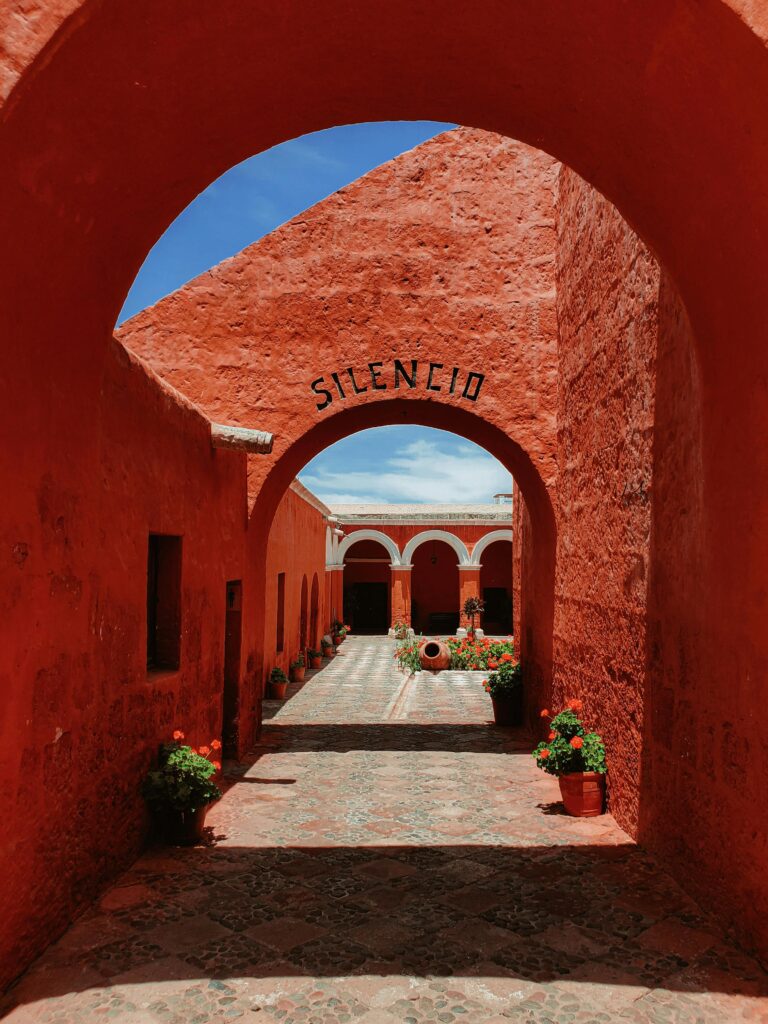 Tranquil monastery courtyard with red walls and potted plants under a sunny archway.